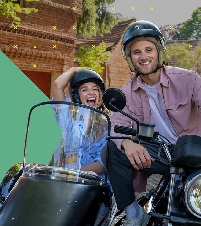Dr. Leon Windscheid sitzt auf einem Motorrad. Neben ihm sitzt Lola Weippert in einem Beiwagen. Beide tragen Helme und lächeln nach vorne. Im Hintergrund ist eine Backsteinwand mit Bäumen zu sehen.