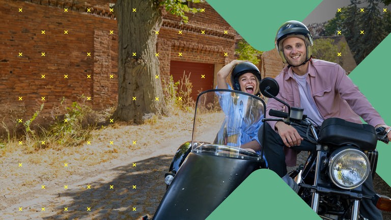 Dr. Leon Windscheid sitzt auf einem Motorrad. Neben ihm sitzt Lola Weippert in einem Beiwagen. Beide tragen Helme und lächeln nach vorne. Im Hintergrund ist eine Backsteinwand mit Bäumen zu sehen.