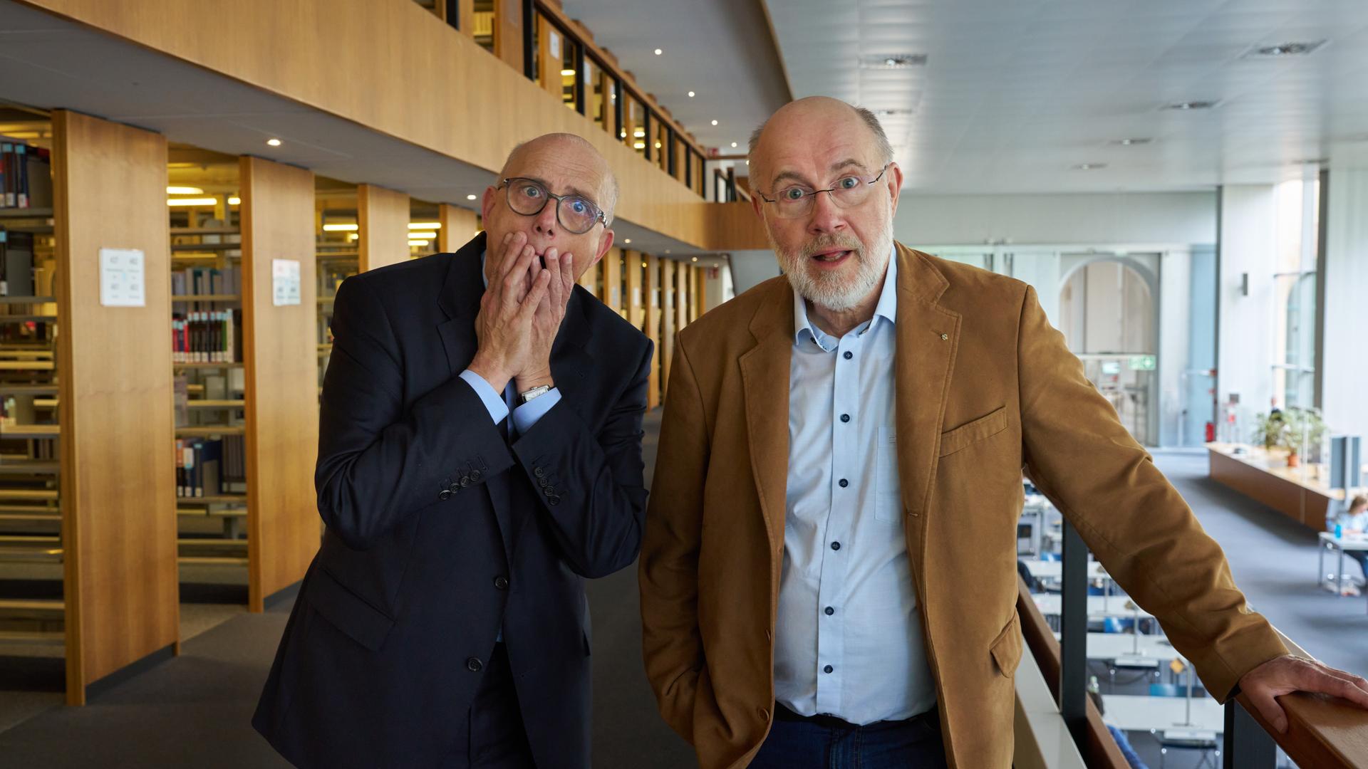 Thomas Schwartz und Harald Lesch stehen im Lesesaal der Bayerischen Staatsbibliothek in München. Sie schauen ungläubig in die Kamera.