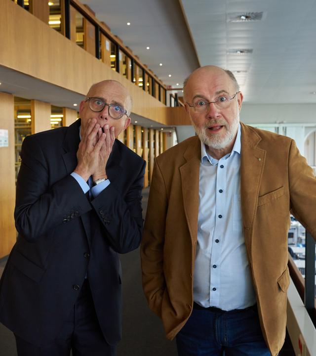 Thomas Schwartz und Harald Lesch stehen im Lesesaal der Bayerischen Staatsbibliothek in München. Sie schauen ungläubig in die Kamera.