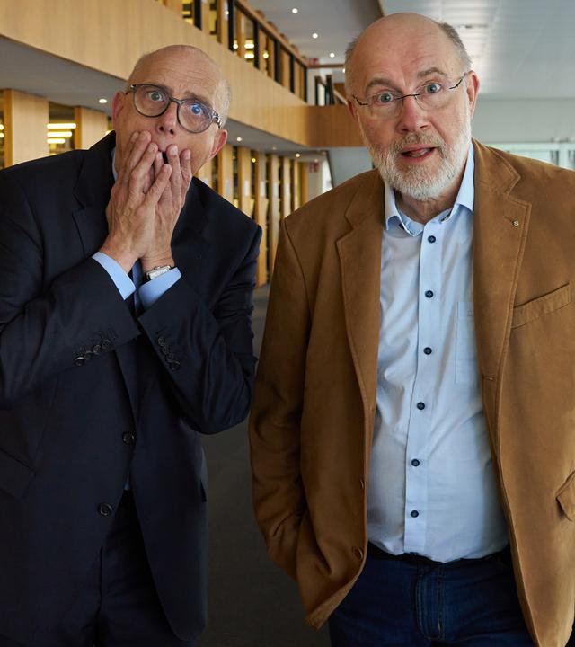 Thomas Schwartz und Harald Lesch stehen im Lesesaal der Bayerischen Staatsbibliothek in München. Sie schauen ungläubig in die Kamera.