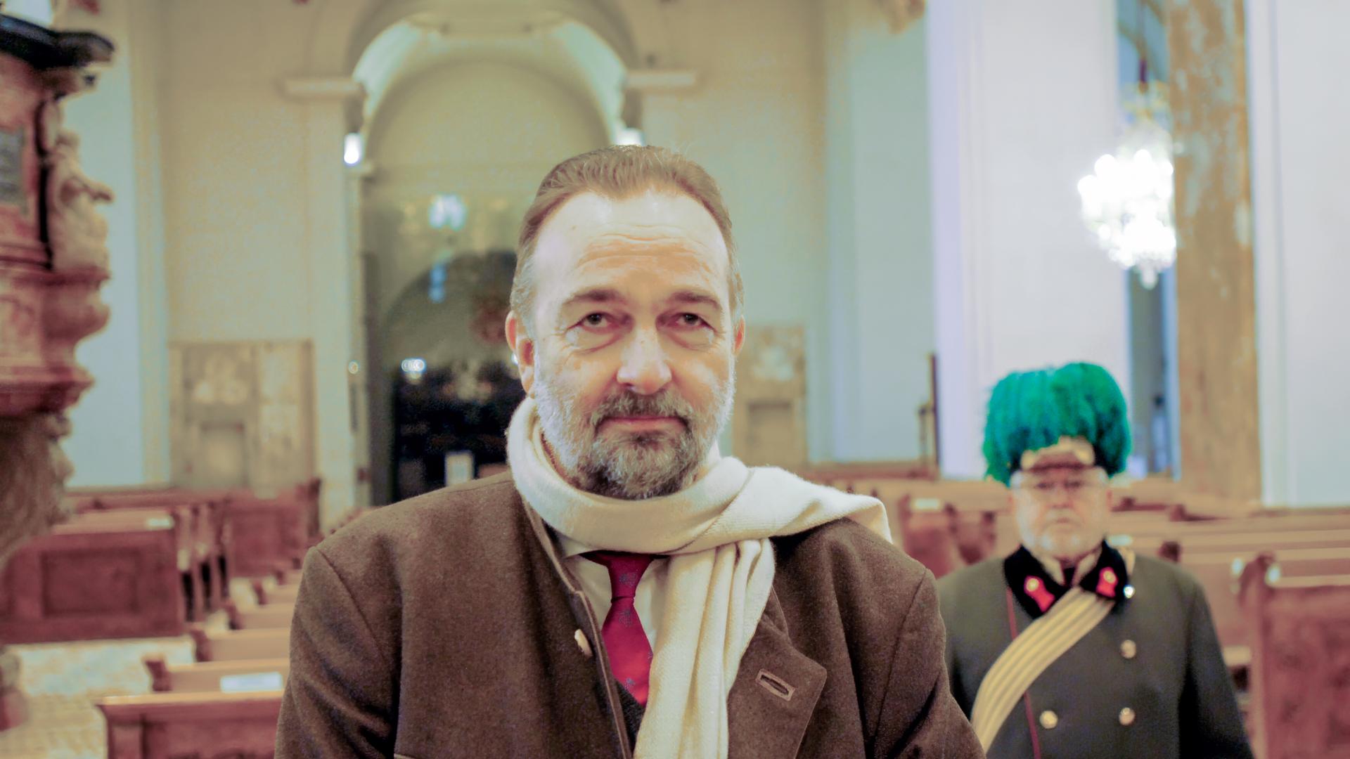 Ein Mann mit einem Bart und einem beigen Schal steht in einer beleuchteten Kirche, hinter ihm ist ein Soldat in historischer Uniform zu sehen.