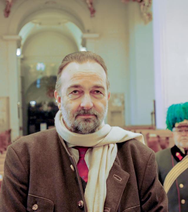 Ein Mann mit einem Bart und einem beigen Schal steht in einer beleuchteten Kirche, hinter ihm ist ein Soldat in historischer Uniform zu sehen.