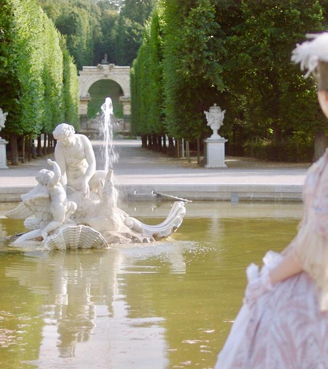 Frau in historischem Kleid blickt auf eine Brunnenfigur in den Gartenanlagen von Schloss Schönbrunn, im Hintergrund ein von Bäumen gesäumter Weg.