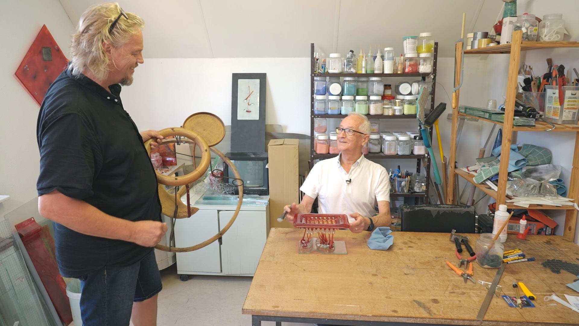 Händler Jos van Katwijk steht mit einem Lauflerngerät für Kinder in der Werkstatt eines Künstlers, der dort an einem Tisch sitzt und arbeitet.