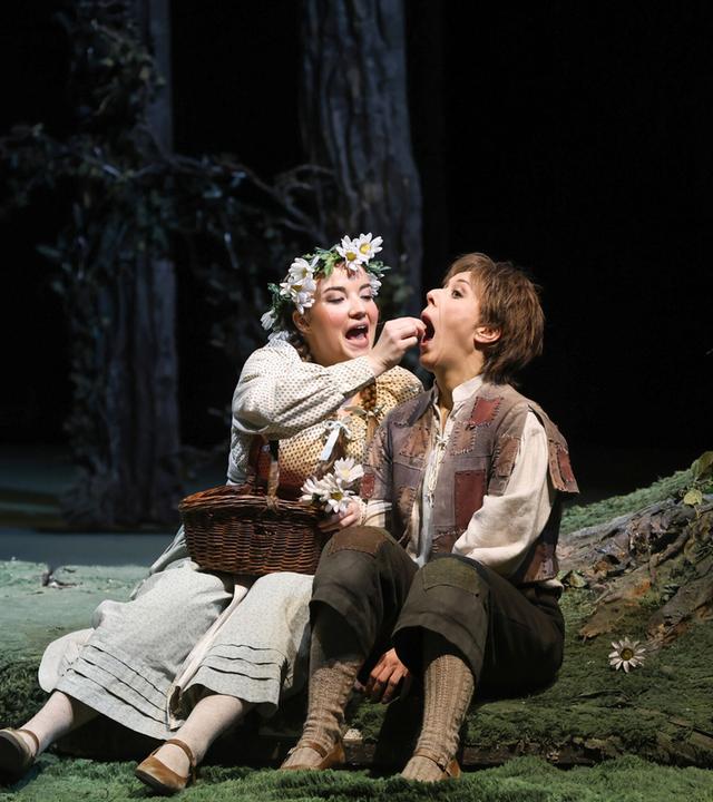 Gretel gibt Hänsel etwas zu essen. Sie sitzen auf einem Waldboden vor Bäumen.