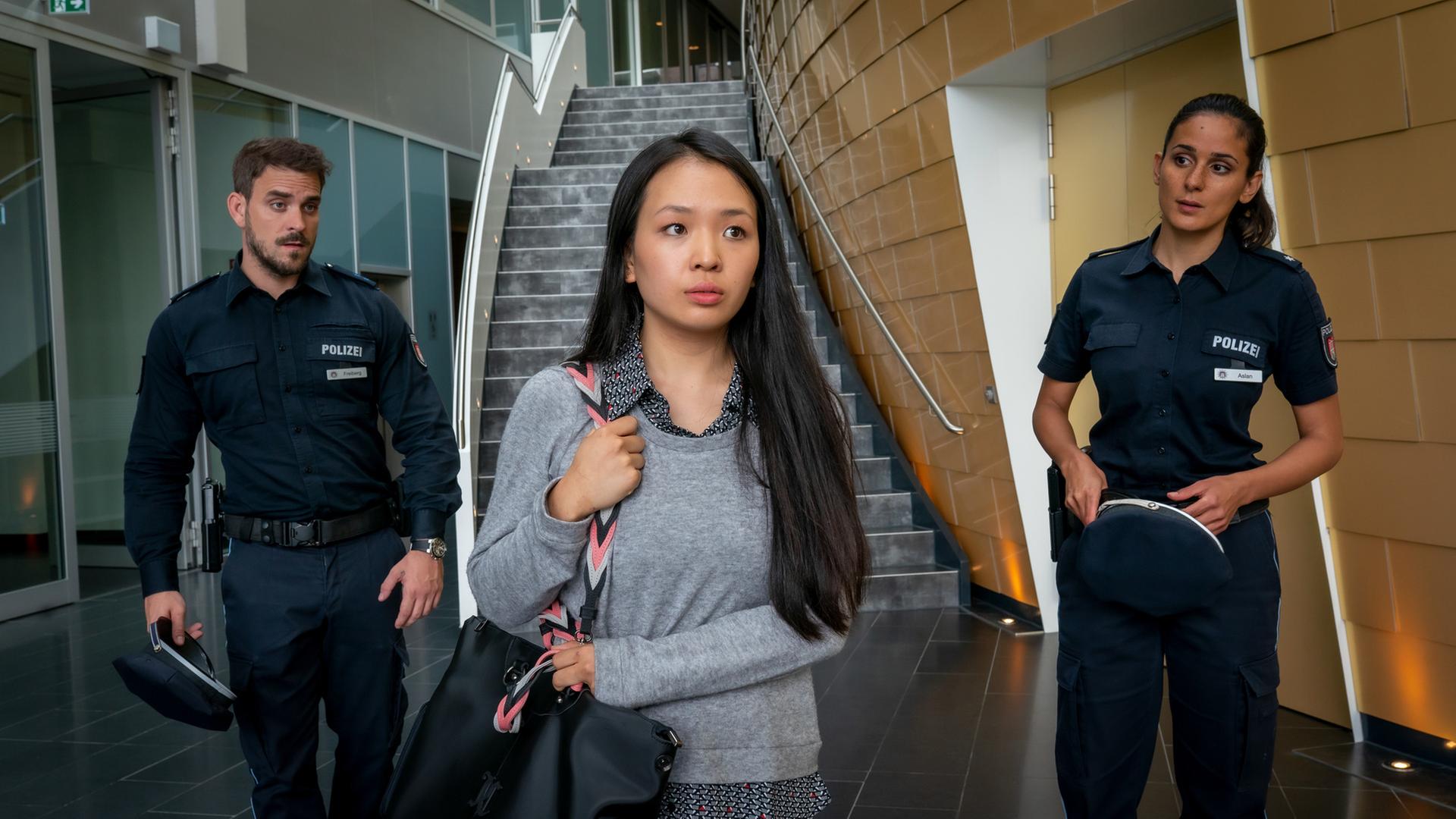 "Notruf Hafenkante - Härtefall": Im Vordergrund steht Gina Weskamp (Andrea Guo). Sie trägt eine Umhängetasche über ihrer rechten Schulter, die sie festhält. Sie blickt nachdenklich ins Leere. Hinter ihr stehen der Polizist Kris Freiberg (Marc Barthel) und seine Kollegin Pinar Aslan (Abi Era). Beide blicken Gina hinterher.