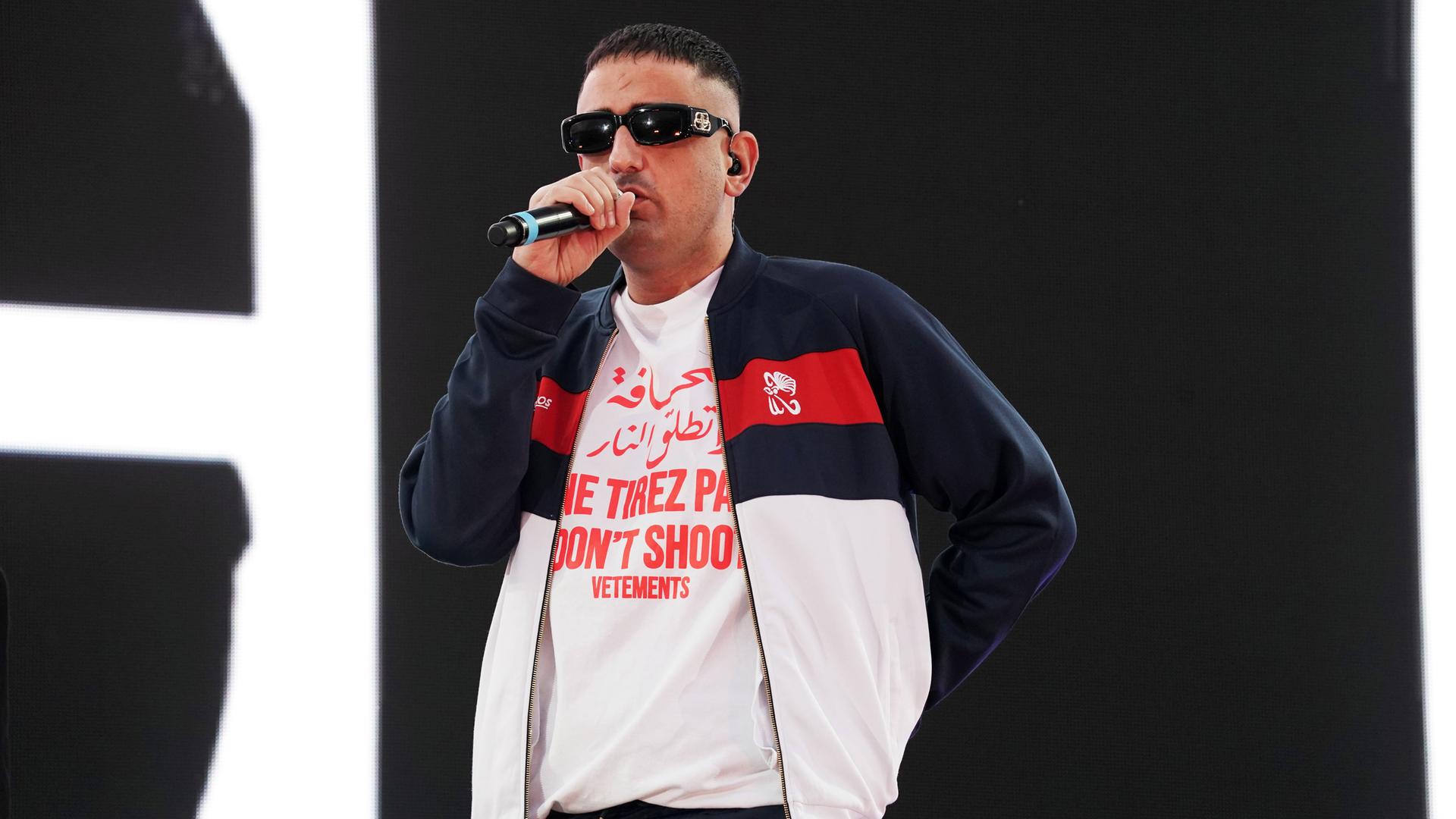 Ein Mann mit Kurzhaarschnitt und Sonnenbrille singt auf einer Bühne in ein Mikro.