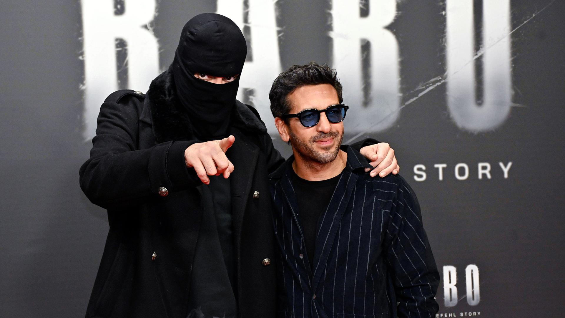 Ein Mann mit schwarzer Skimaske und ein zweiter stehen vor Filmwerbung.