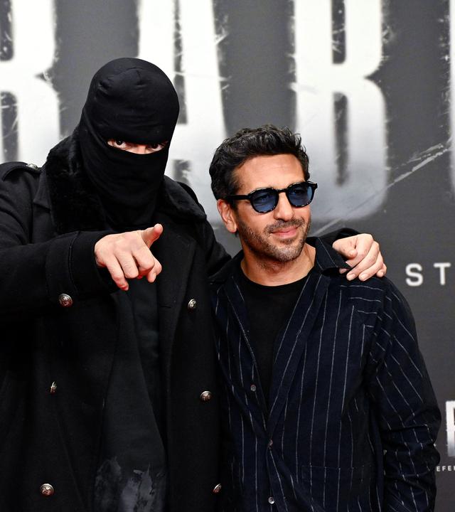 Ein Mann mit schwarzer Skimaske und ein zweiter stehen vor Filmwerbung.