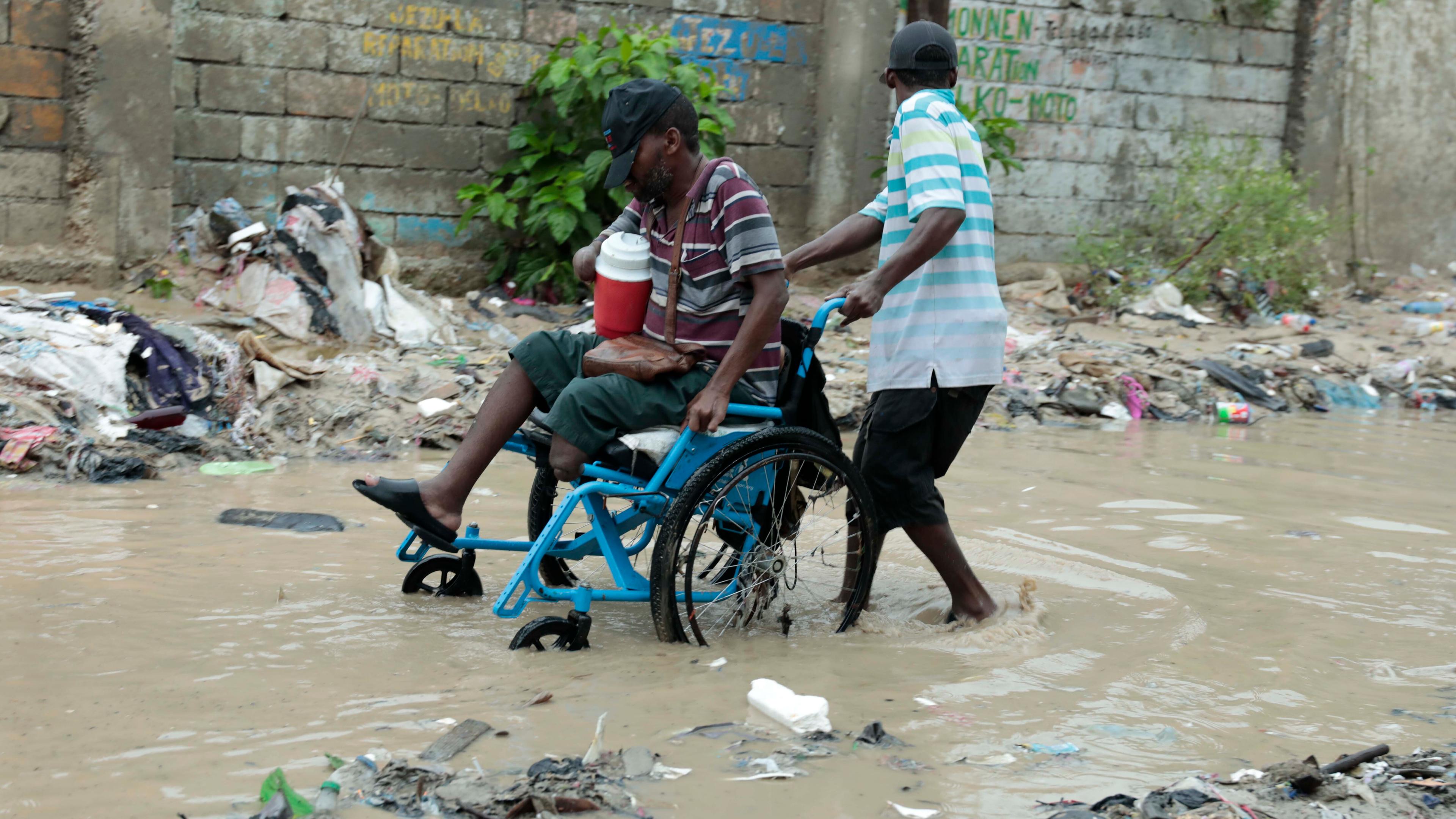 Einem Mann im Rollstuhl wird geholfen, sich einen Weg über eine überflutete Straße nach starkem Regen zu bahnen.