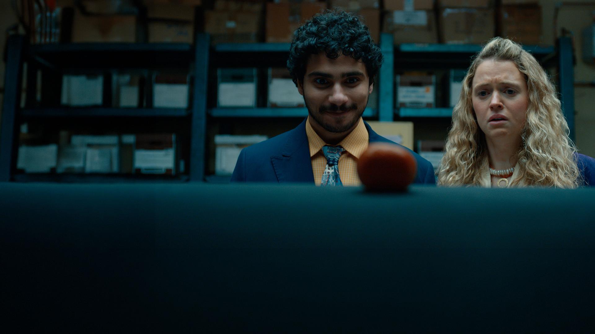 "Extraordinary - Hallo Fremder": Carrie (Sofia Oxenham) und Kash (Bilal Hasna) stehen nebeneinander in einem Lagerraum mit blauen Metallregalen und zahlreichen Kartons. Beide blicken konzentriert auf einen runden, rot-orangefarbenen Gegenstand im Vordergrund, wobei Kash zufrieden wirkt, während Carries Gesichtsausdruck deutlich besorgt erscheint.