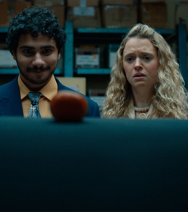"Extraordinary - Hallo Fremder": Carrie (Sofia Oxenham) und Kash (Bilal Hasna) stehen nebeneinander in einem Lagerraum mit blauen Metallregalen und zahlreichen Kartons. Beide blicken konzentriert auf einen runden, rot-orangefarbenen Gegenstand im Vordergrund, wobei Kash zufrieden wirkt, während Carries Gesichtsausdruck deutlich besorgt erscheint.