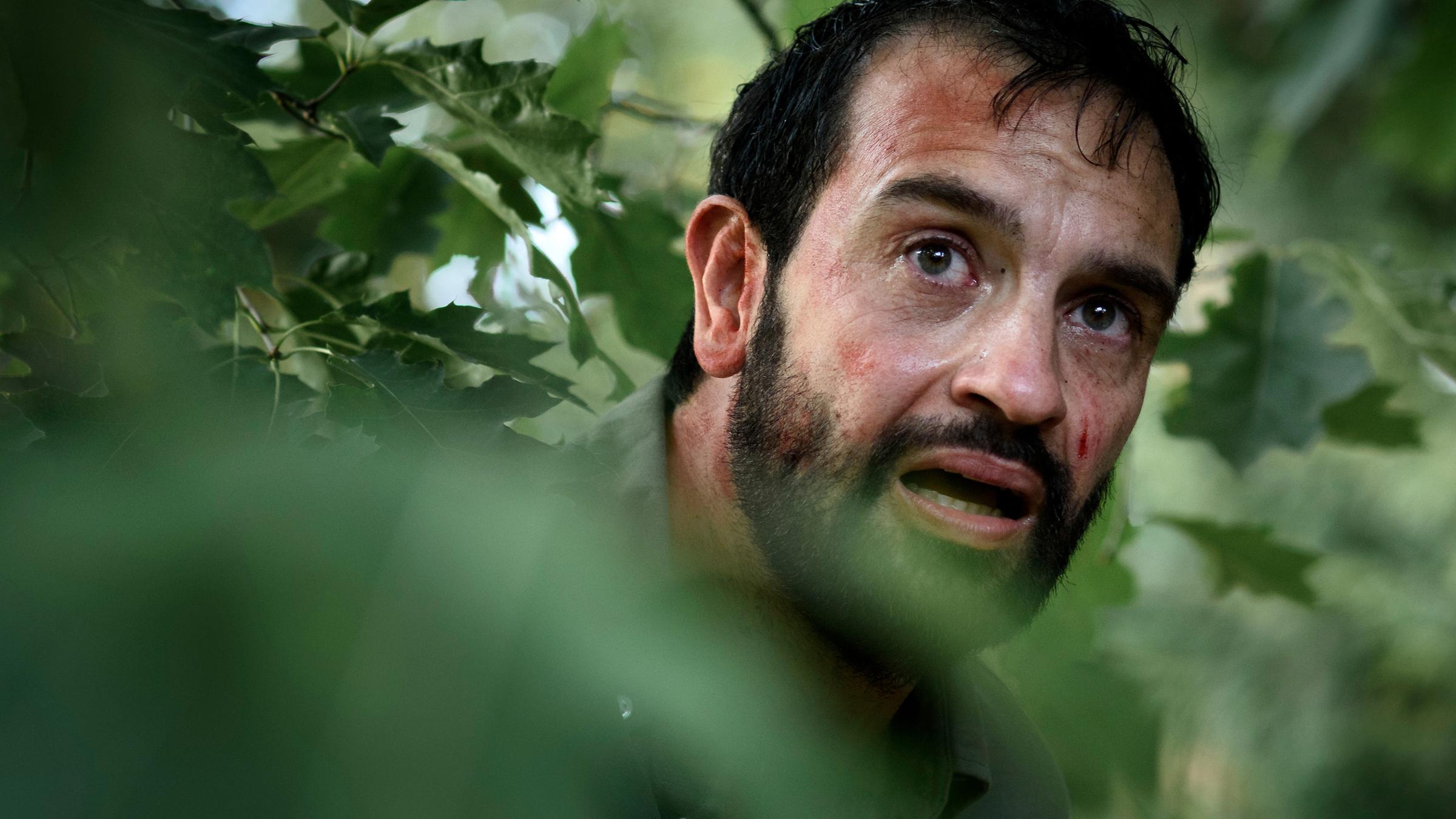 Hans (Jeroen Spitzenberger) ist auf der Flucht und versteckt sich in einem Gebüsch. Sein Gesicht ist verschwitzt und blutverschmiert, wobei er verängstigt mit offenem Mund gerade aus dem Gestrüpp herausblickt.
