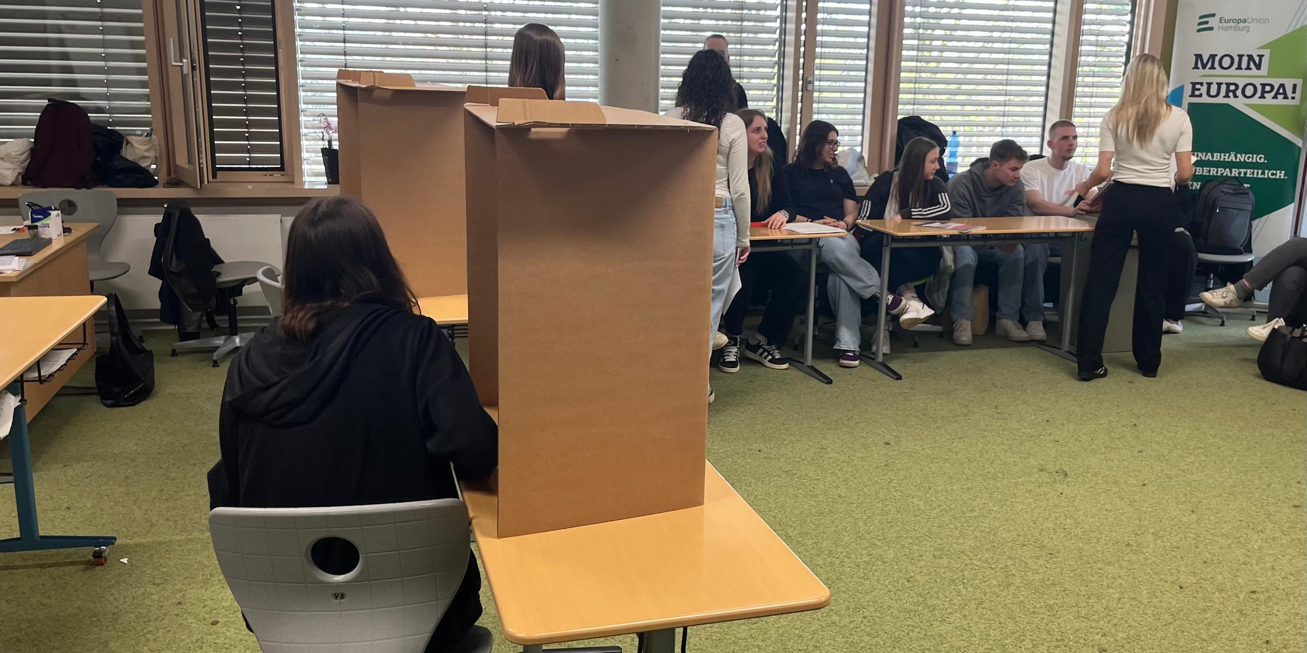 In einem Klassenraum steht eine Wahlkabine. Eine Schülerin gibt ihre Stimme ab.