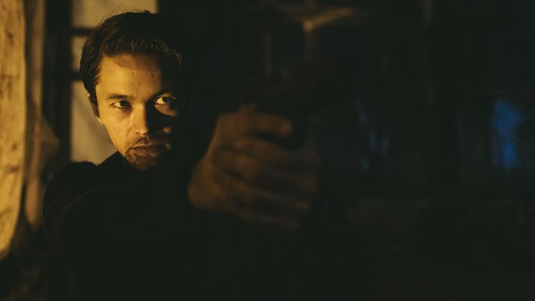 "Hamilton - Undercover in Stockholm": Agent Hamilton (Jakob Oftebro) steht im Halbdunkel mit einer Pistole im Anschlag und zielt rechts aus dem Bild.