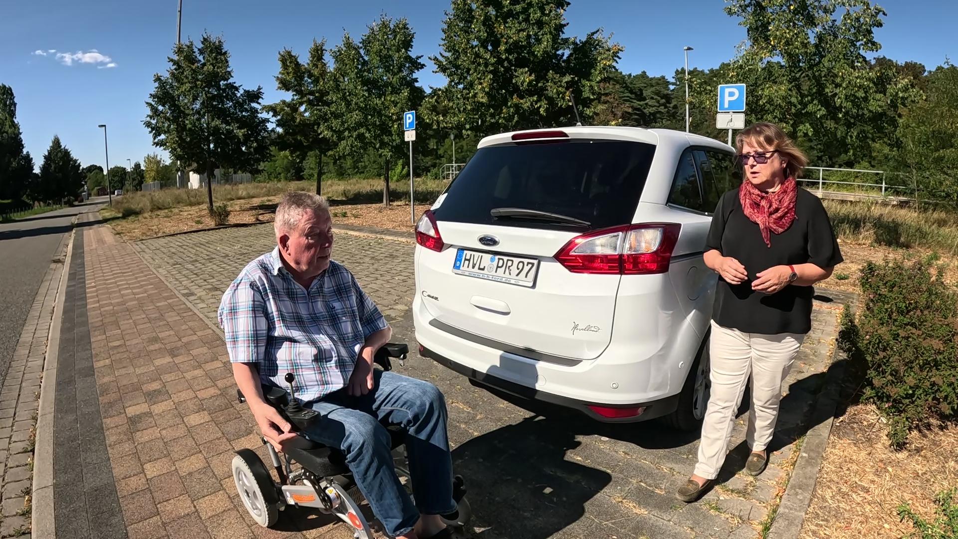 Ein Mann im Rollstuhl hinter einem PKW auf einem Behindertenparkplatz, neben der Beifahrertür kommt eine Frau auf den Rollstuhlfahrer zu