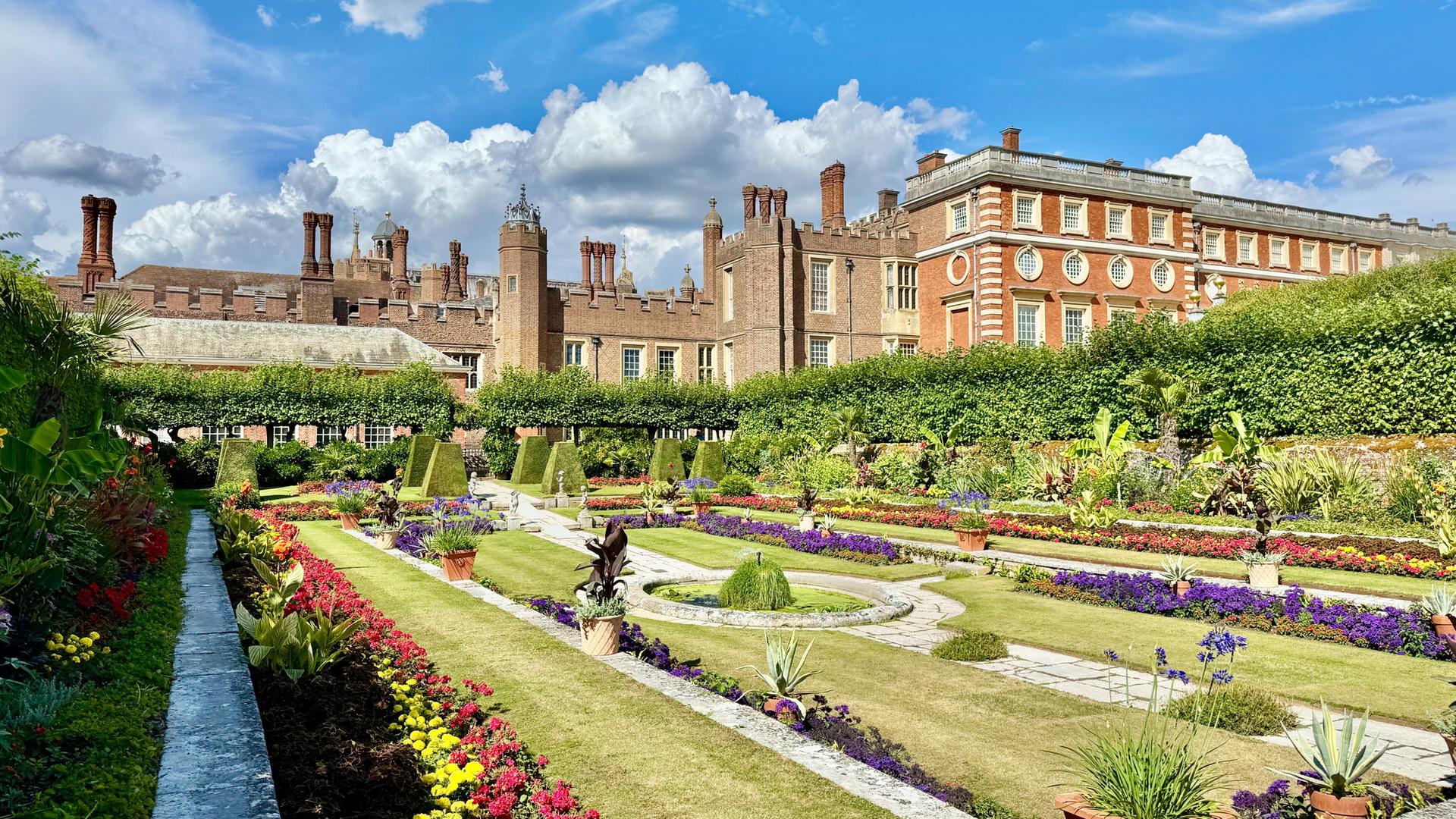 Das Bild zeigt die königlichen Gärten von Hampton Court Palace. Im Vordergrund sind prachtvolle barocke Gärten zu sehen, die mit bunten Blumenbeeten in verschiedenen Formen und Farben gestaltet sind. Ein schmaler Weg aus Stein zieht sich durch die Gartenanlage.   Im Hintergrund erhebt sich das Gebäude des Hampton Court Palace, das aus roten Ziegelsteinen und grauem Stein besteht. Es ist von hohen, grünen Hecken umgeben, die eine klare Abgrenzung zu den Gartenbereichen bilden. Die Fassade des Palastes zeigt verschiedene architektonische Details, darunter runde Fenster und steinerne Verzierungen.   Der Himmel ist blau mit einigen weißen, wolkigen Strukturen, die für eine helle und freundliche Atmosphäre sorgen. Das Bild vermittelt einen Eindruck von harmonischer Gartenkunst, Geschichte und künstlerischem Erbe.