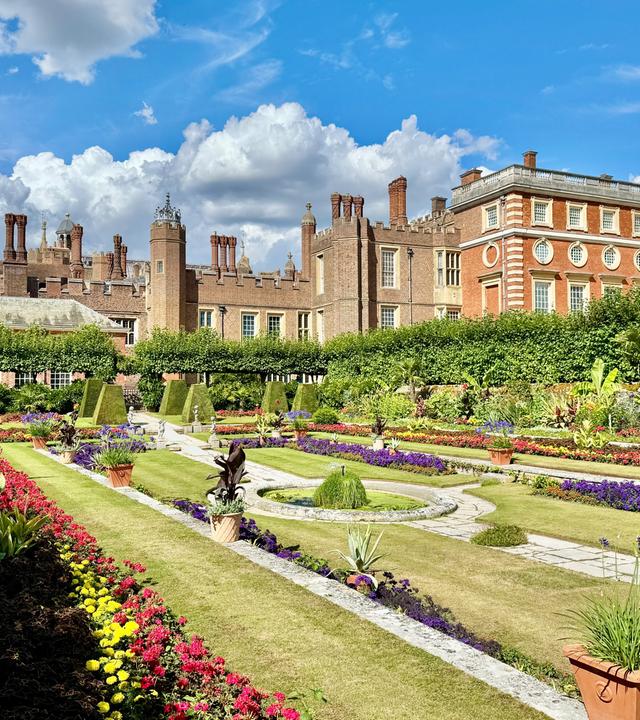 Das Bild zeigt die königlichen Gärten von Hampton Court Palace. Im Vordergrund sind prachtvolle barocke Gärten zu sehen, die mit bunten Blumenbeeten in verschiedenen Formen und Farben gestaltet sind. Ein schmaler Weg aus Stein zieht sich durch die Gartenanlage.   Im Hintergrund erhebt sich das Gebäude des Hampton Court Palace, das aus roten Ziegelsteinen und grauem Stein besteht. Es ist von hohen, grünen Hecken umgeben, die eine klare Abgrenzung zu den Gartenbereichen bilden. Die Fassade des Palastes zeigt verschiedene architektonische Details, darunter runde Fenster und steinerne Verzierungen.   Der Himmel ist blau mit einigen weißen, wolkigen Strukturen, die für eine helle und freundliche Atmosphäre sorgen. Das Bild vermittelt einen Eindruck von harmonischer Gartenkunst, Geschichte und künstlerischem Erbe.