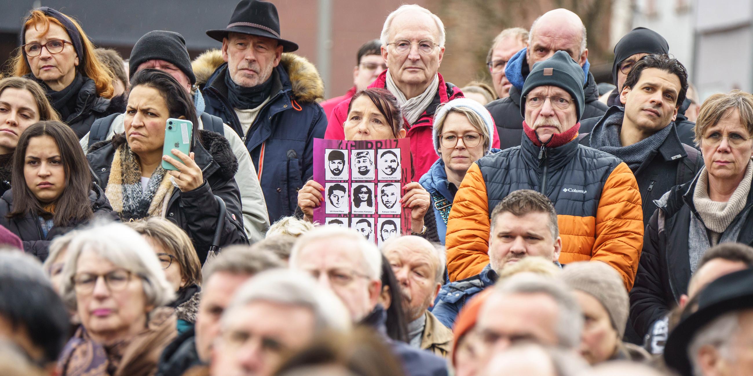 Eine Frau (M) hält ein Plakat mit Abbildungen der Opfer in der Hand bei einer Gedenkstunde auf dem Marktplatz, bei der an die neun Opfer des rassistischen Anschlags in Hanau vor drei Jahren erinnert wird.