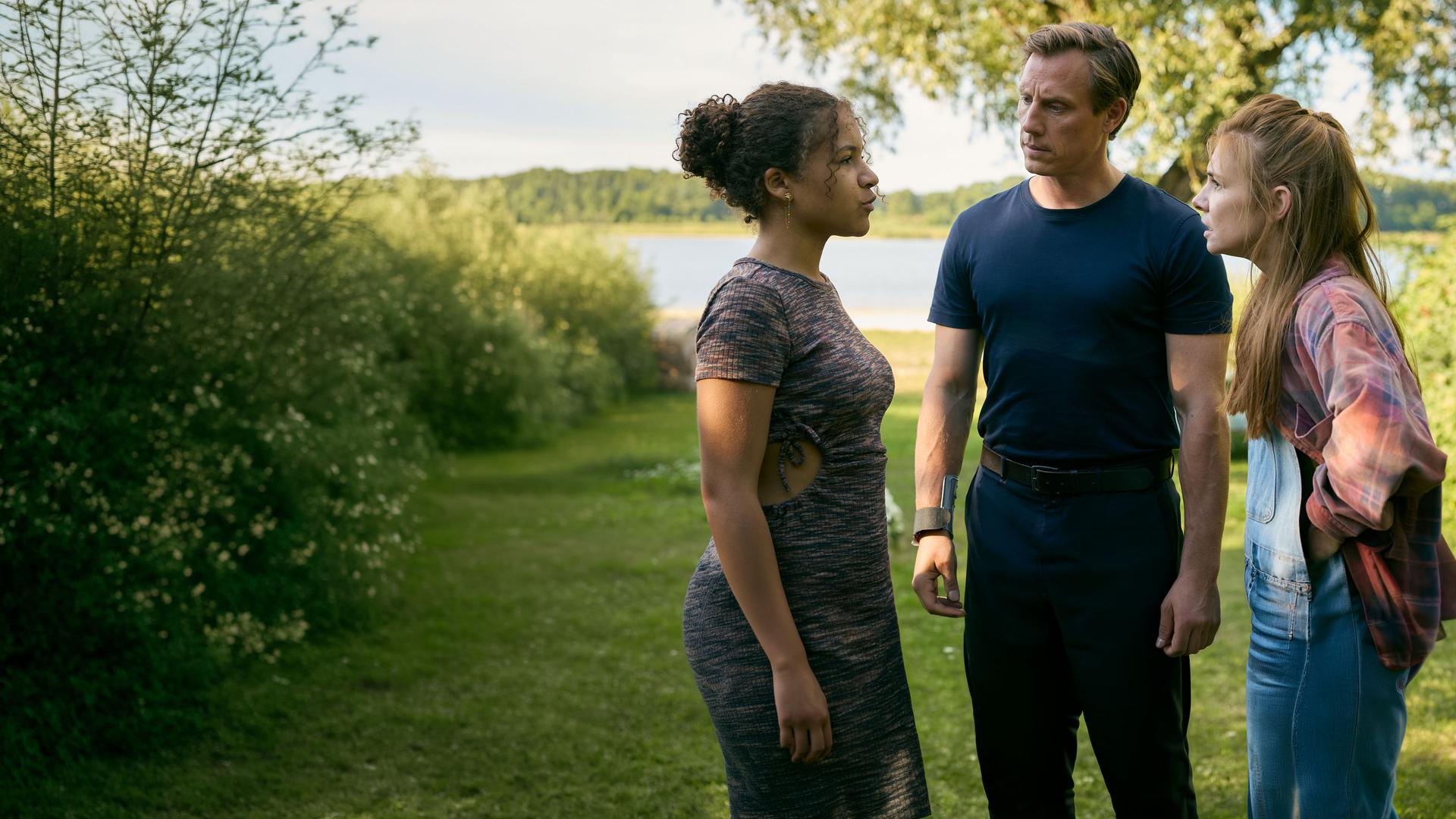 "Dr. Nice - Hand aufs Herz": In einer grün mit Sträuchern bewachsenen Landschaft steht Charlie (Josefine Preuß) wütend Lea (Maj Borchardt) gegenüber. Leas "Vater" Moritz Neiss (Patrick Kalupa) steht zwischen den beiden und schaut Lea fragend an. In der Ferne ist ein kleiner See zu sehen.