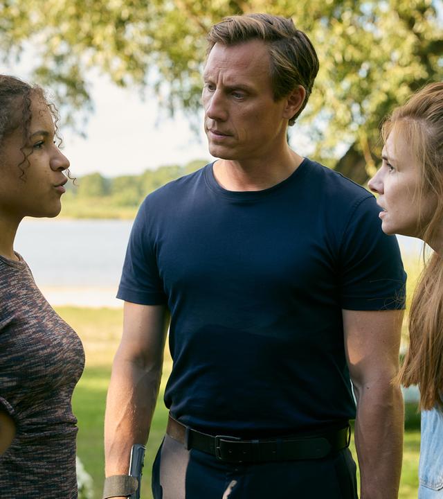 "Dr. Nice - Hand aufs Herz": In einer grün mit Sträuchern bewachsenen Landschaft steht Charlie (Josefine Preuß) wütend Lea (Maj Borchardt) gegenüber. Leas "Vater" Moritz Neiss (Patrick Kalupa) steht zwischen den beiden und schaut Lea fragend an. In der Ferne ist ein kleiner See zu sehen.