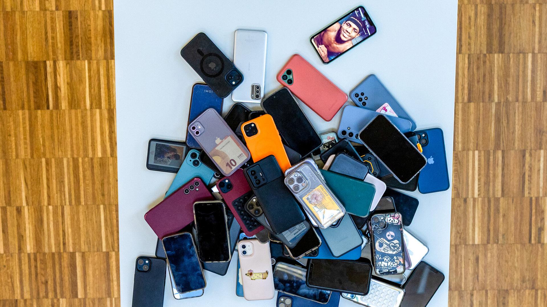 Das Bild zeigt einen Haufen von Mobiltelefonen, die auf einem weißen Tisch abgeladen sind. Die Telefone sind in verschiedenen Farben und Modellen vorhanden, darunter schwarze, graue, blaue, grüne, rote und lilafarbene Geräte. Einige Handys sind ohne Hülle, während andere mit unterschiedlich gestalteten Schutzhüllen versehen sind. Auf einem der Handys ist das Bild eines Menschen zu sehen, möglicherweise ein Selfie. Der Tisch steht auf einem Boden, der aus Holzplanken besteht und eine warme, braune Farbgebung hat. Die Szene wirkt chaotisch und verdeutlicht die große Anzahl an Handys, die hier gesammelt wurden.