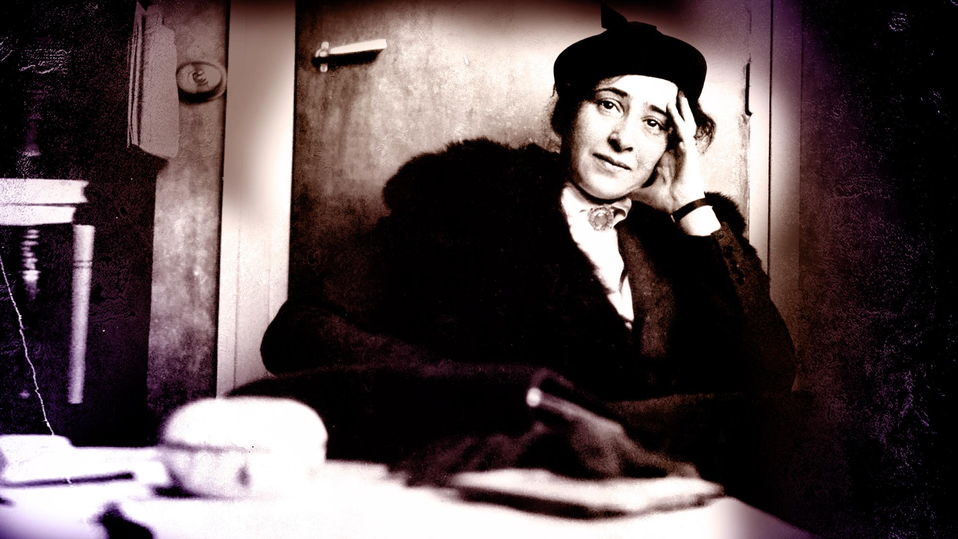 Hannah Arendt sitzt auf einem Sessel bei einem Fernsehinterview. Sie trägt ein schwarzes Kostüm und ihr Ellenbogen stützt sie auf der Armlehne auf. IN der Hand hält sie eine ZIgarette. Das Bild ist schwarz weiß. 