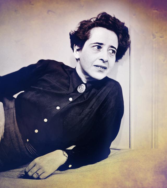 Hannah Arendt sitzt auf einem Sessel bei einem Fernsehinterview. Sie trägt ein schwarzes Kostüm und ihr Ellenbogen stützt sie auf der Armlehne auf. IN der Hand hält sie eine ZIgarette. Das Bild ist schwarz weiß. 