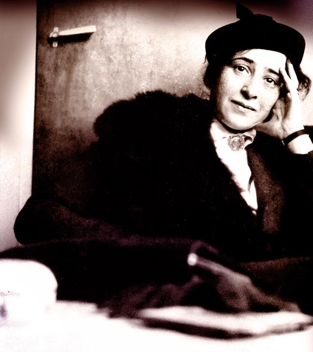 Hannah Arendt sitzt auf einem Sessel bei einem Fernsehinterview. Sie trägt ein schwarzes Kostüm und ihr Ellenbogen stützt sie auf der Armlehne auf. IN der Hand hält sie eine ZIgarette. Das Bild ist schwarz weiß. 