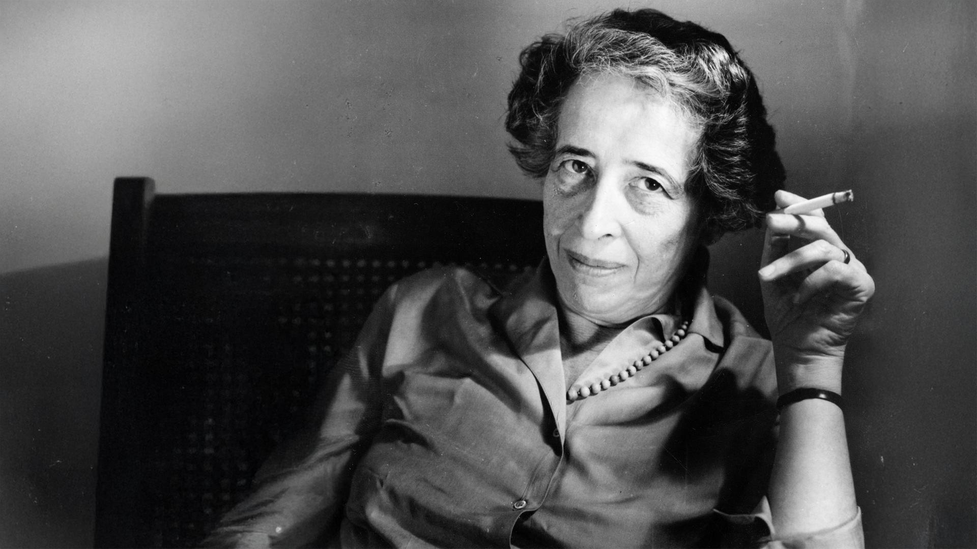 Altes Schwarz-Weiß-bild von einer Frau mittleren Alters, die sich in einem Stuhl zurücklehnt und eine rauchende Zigarette zwischen zwei Fingern hält.