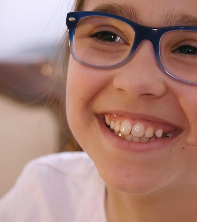 Auf diesem Bild ist fast nur Hannahs Gesicht zu sehen sowie ein Teil ihrer Schultern. Sie lacht sehr offen und fröhlich, trägt eine Brille mit blauem Gestell und eine weiße Bluse.