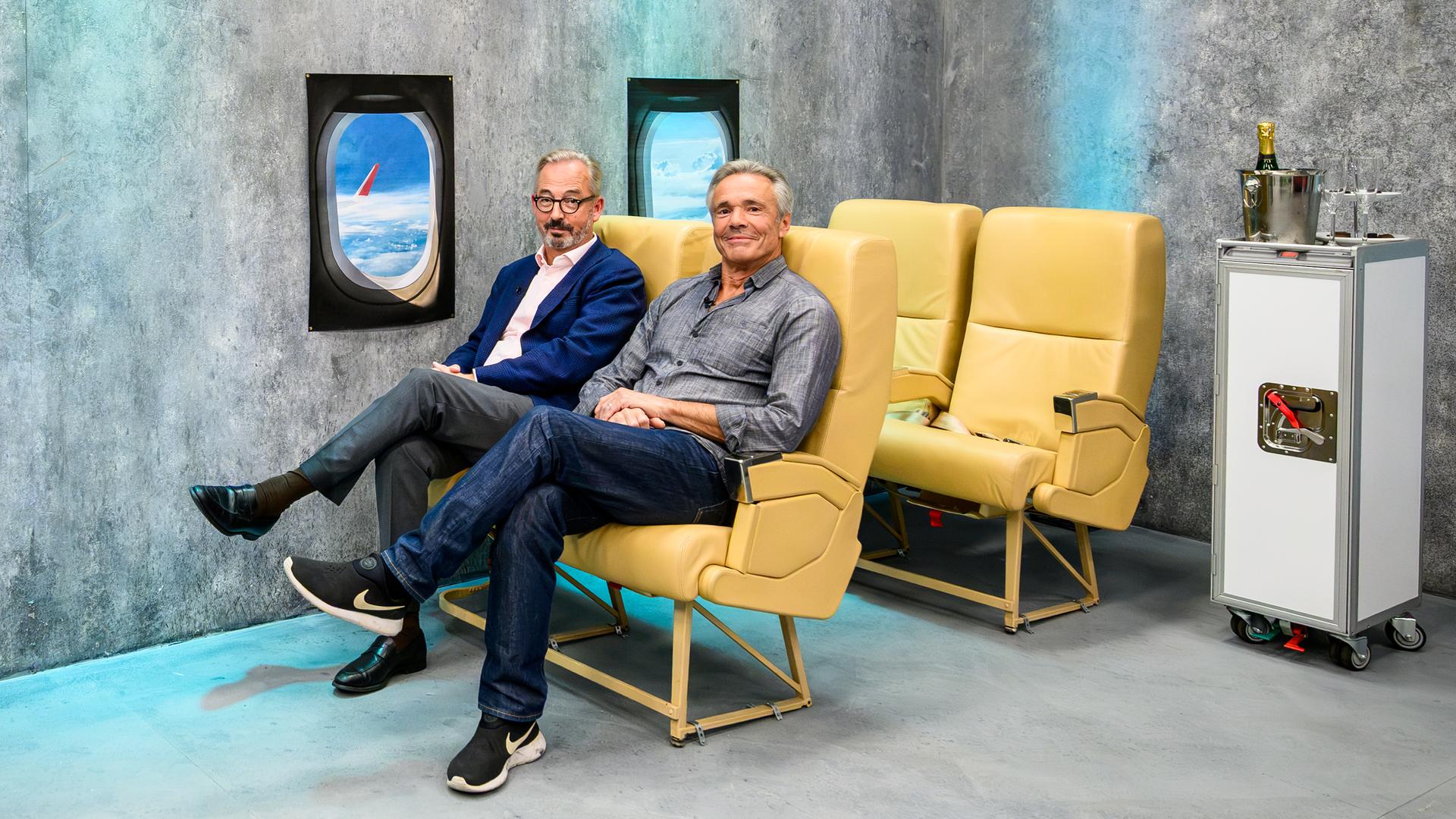 Jan Fleischhauer und Hannes Jaenicke sitzen nebeneinander auf gelben Flugzeugsitzen vor einer grauen Studiowand. Fleischhauer trägt ein blaues Sakko und eine graue Hose, Jaenicke ein graues Hemd, Jeans und schwarze Sportschuhe. An der Wand sind zwei nachgebildete Flugzeugfenster mit Wolken und einem Flugzeugflügel zu sehen. Rechts steht ein Servierwagen mit einer Flasche im Kühler und mehreren Gläsern.
