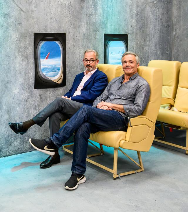 Jan Fleischhauer und Hannes Jaenicke sitzen nebeneinander auf gelben Flugzeugsitzen vor einer grauen Studiowand. Fleischhauer trägt ein blaues Sakko und eine graue Hose, Jaenicke ein graues Hemd, Jeans und schwarze Sportschuhe. An der Wand sind zwei nachgebildete Flugzeugfenster mit Wolken und einem Flugzeugflügel zu sehen. Rechts steht ein Servierwagen mit einer Flasche im Kühler und mehreren Gläsern.