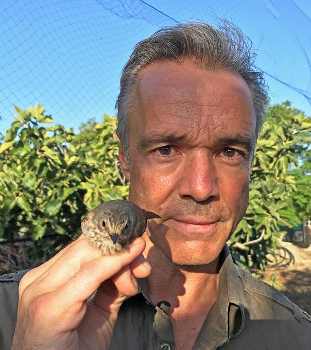 Hannes Jaenicke hält einen Singvogel in der rechten Hand. Im Hintergrund sieht man Bäume und blauen Himmel.