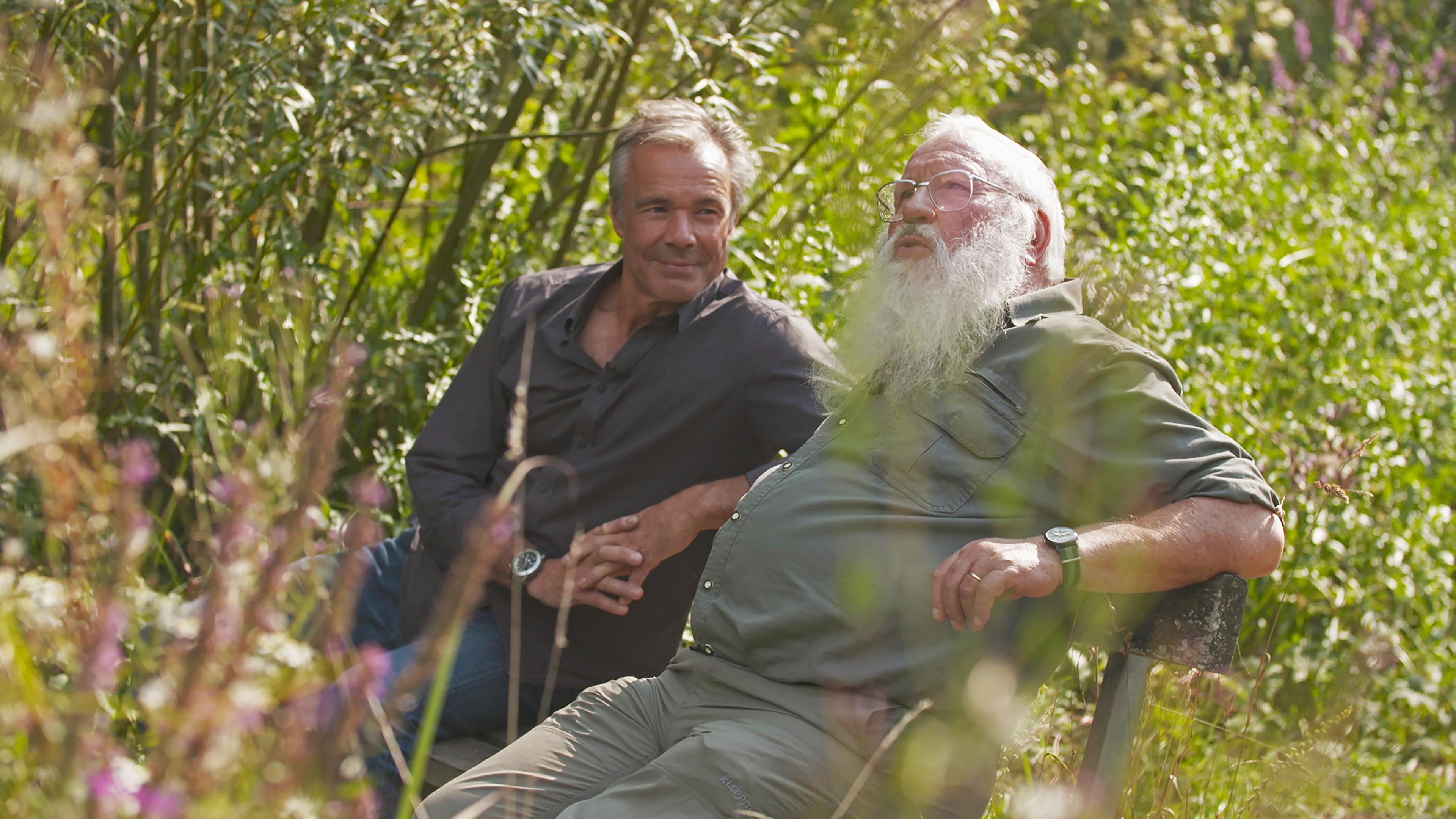 Hannes Jaenicke sitzt neben dem Vogelexperten Prof. Peter Berthold auf einer Bank im Grünen.