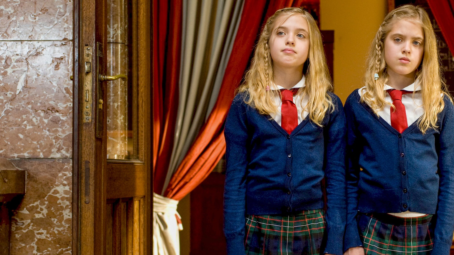 Hanni und Nanni stehen in Schuluniform nebeneinander zwischen zwei offenen Vorhängen und schauen missmutig.