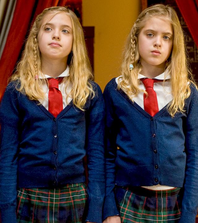 Hanni und Nanni stehen in Schuluniform nebeneinander zwischen zwei offenen Vorhängen und schauen missmutig.