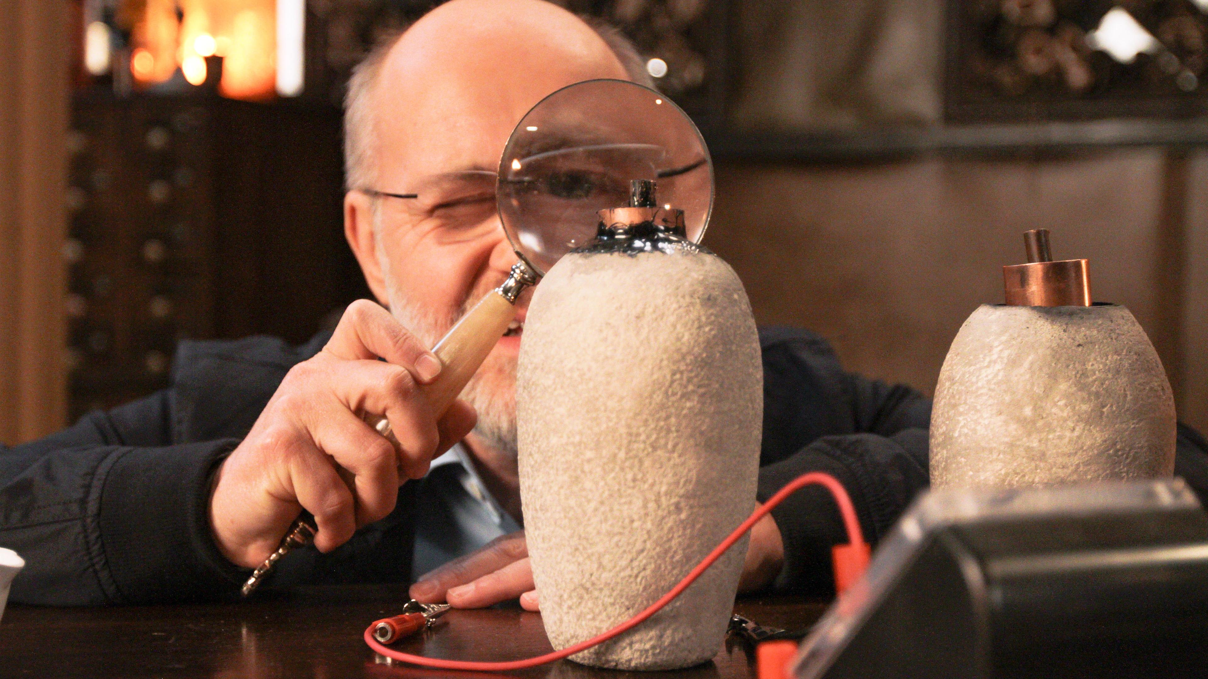 Harald Lesch betrachtet durch eine große Lupe zwei Nachbauten einer "Bagdad-Batterie".