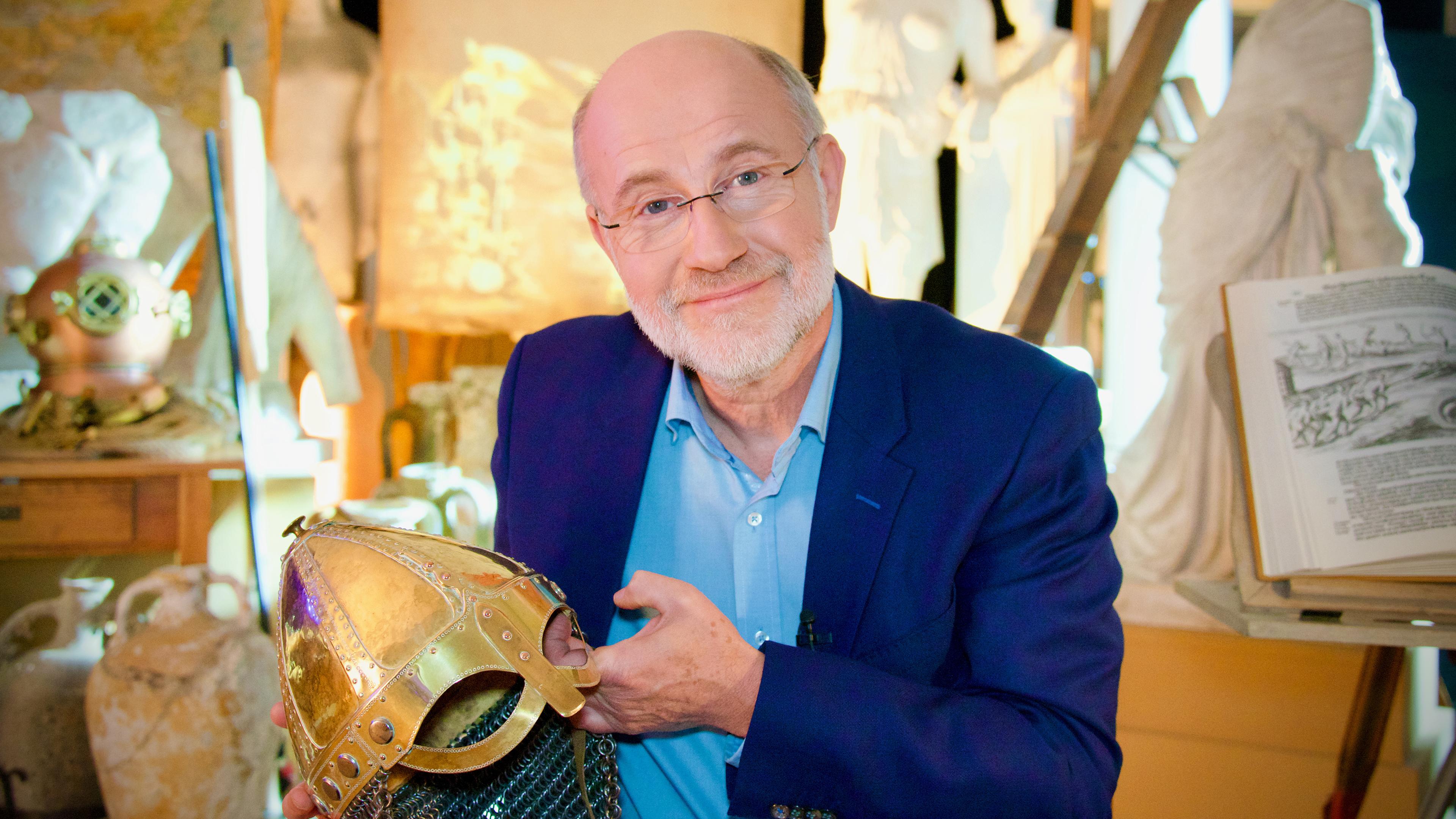 Der Moderator Harald Lesch sitzt in einem Raum mit diversen Nachbildungen antiker Gegenstände und Figuren. In seiner Hand hält er die Nachbildung eines goldfarbenen Kriegshelmes.