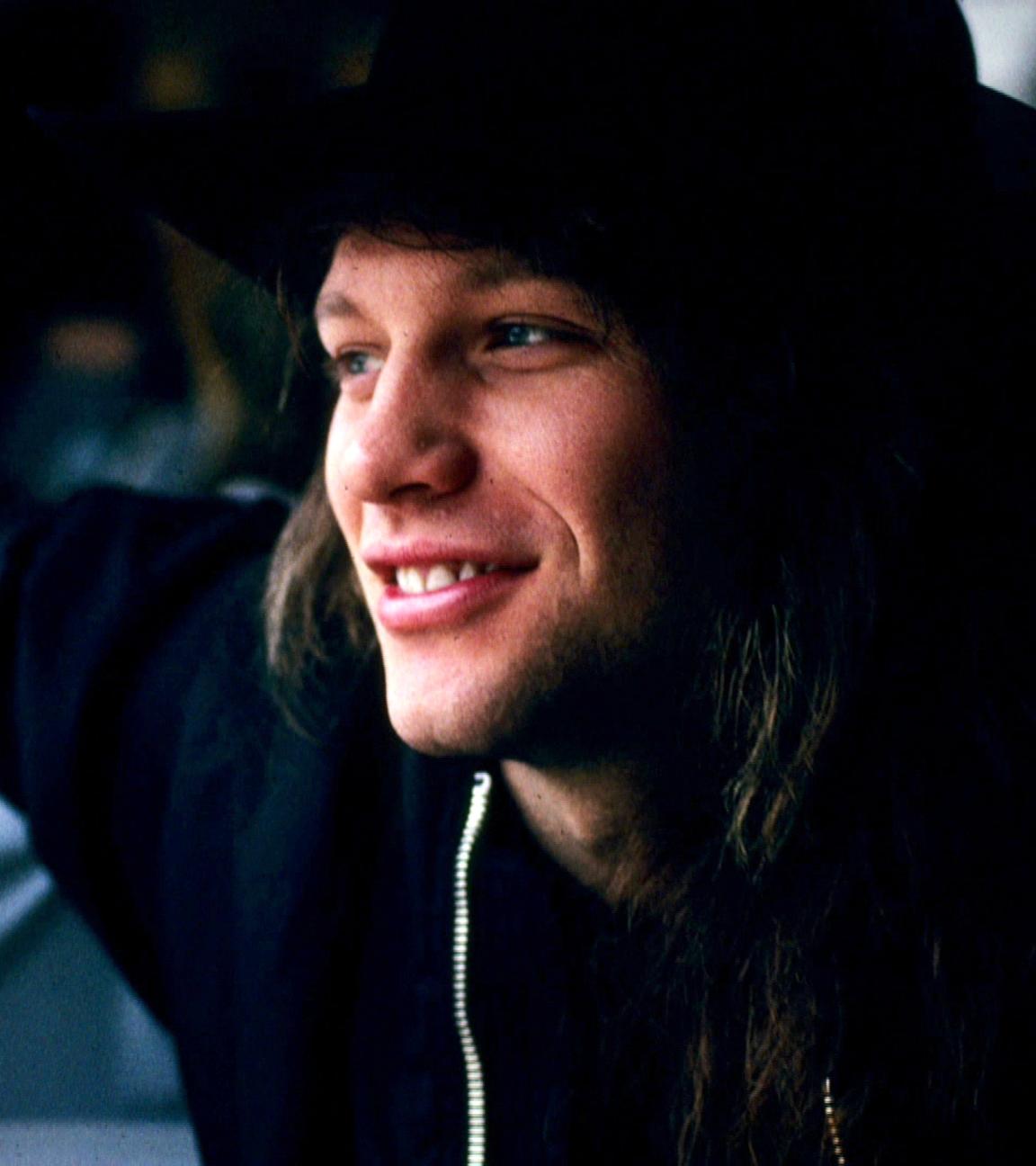 Jon Bon Jovi trägt einen schwarzen Hut und lächelt. Er hat seinen rechten Arm abgestützt und schaut zur Seite.