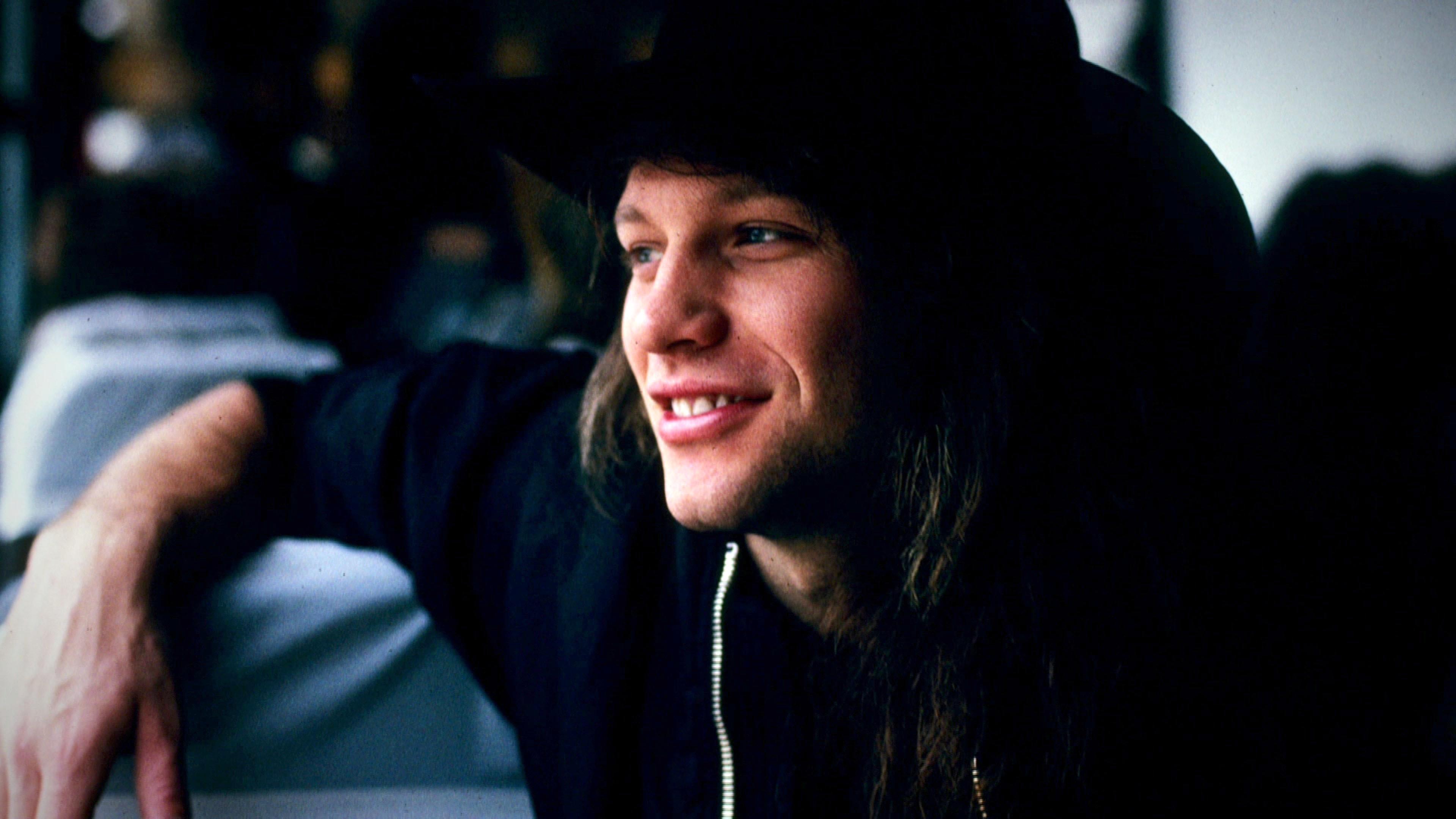 Jon Bon Jovi trägt einen schwarzen Hut und lächelt. Er hat seinen rechten Arm abgestützt und schaut zur Seite.