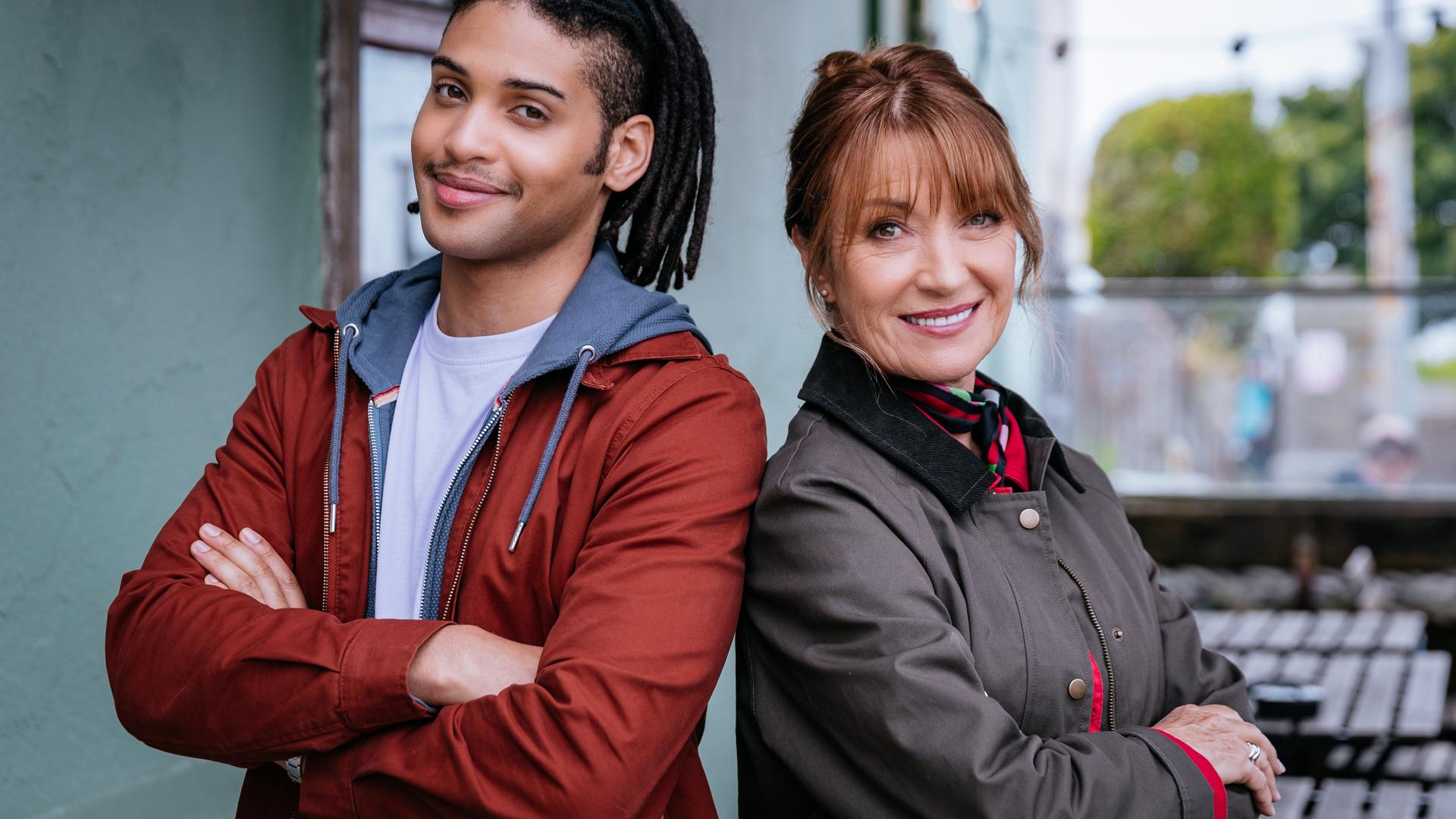 "Harry Wild - Mörderjagd in Dublin": Fergus (Rohan Nedd) und Harry (Jane Seymour) in Nahaufnahme. Sie stehen nebeneinander, die Arme vor sich verschränkt, und lächeln in die Kamera.