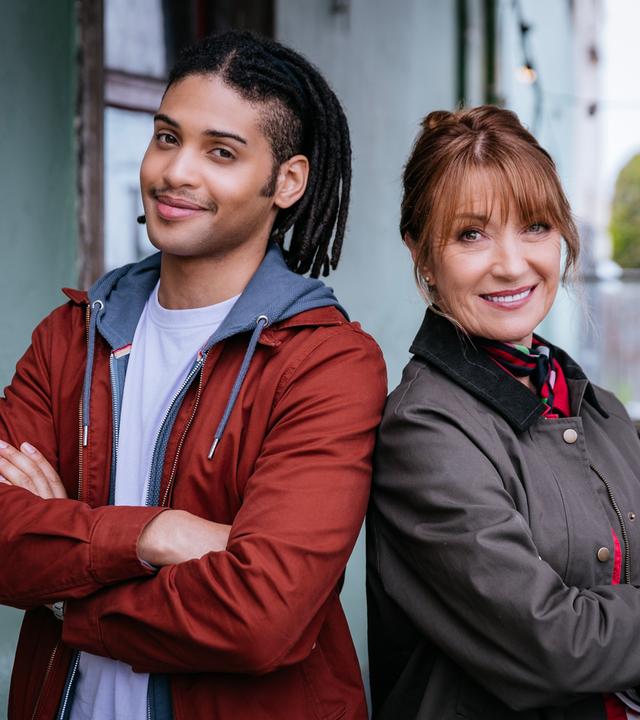 "Harry Wild - Mörderjagd in Dublin": Fergus (Rohan Nedd) und Harry (Jane Seymour) in Nahaufnahme. Sie stehen nebeneinander, die Arme vor sich verschränkt, und lächeln in die Kamera.