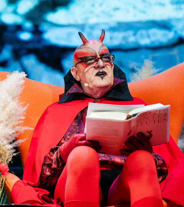 Ein Mann im Mephisto-Kostüm sitzt auf einem orangenen Sessel und hält das Buch "Unterleuten" von Juli Zeh in den Händen