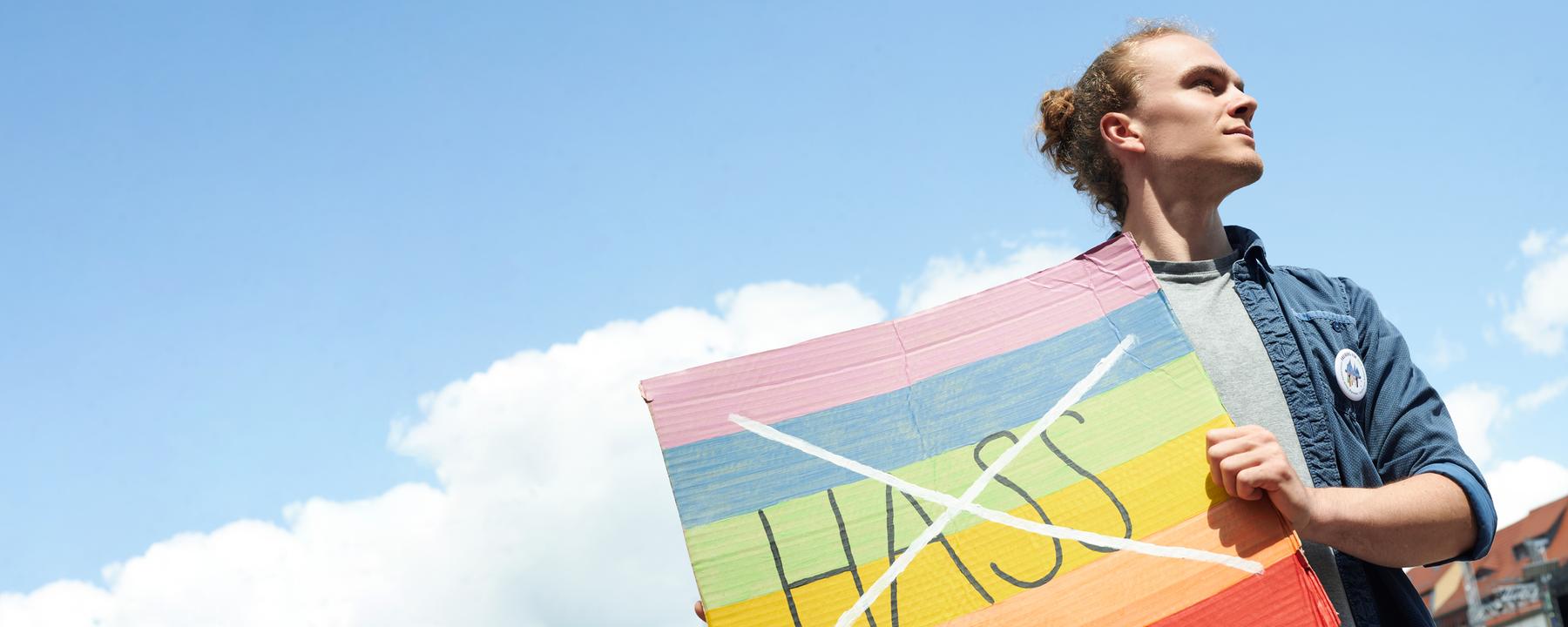 Florian hält ein buntes Schild vor seinen Körper. Auf dem Schild ist das Wort "Hass" durchgestrichen. Er blickt zur Seite.