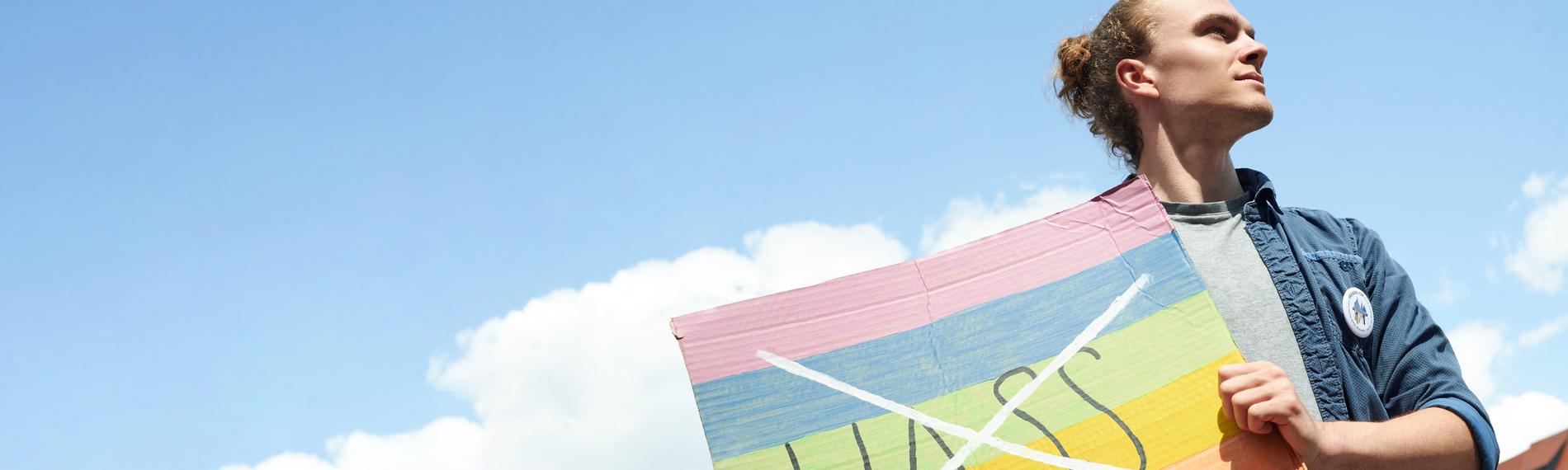 Florian hält ein buntes Schild vor seinen Körper. Auf dem Schild ist das Wort "Hass" durchgestrichen. Er blickt zur Seite.