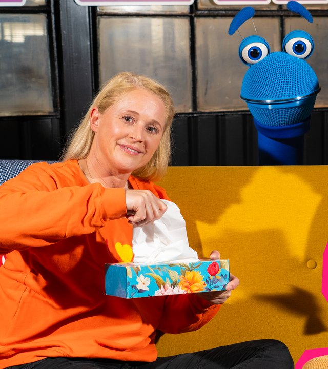 Singa und Simon sagt sitzen auf dem Sofa und schauen leicht kränklich. Singa hält ein Taschentuch in der Hand und Simon eine Tasse. Hinter dem Sofa schaut das SingDing hervor.