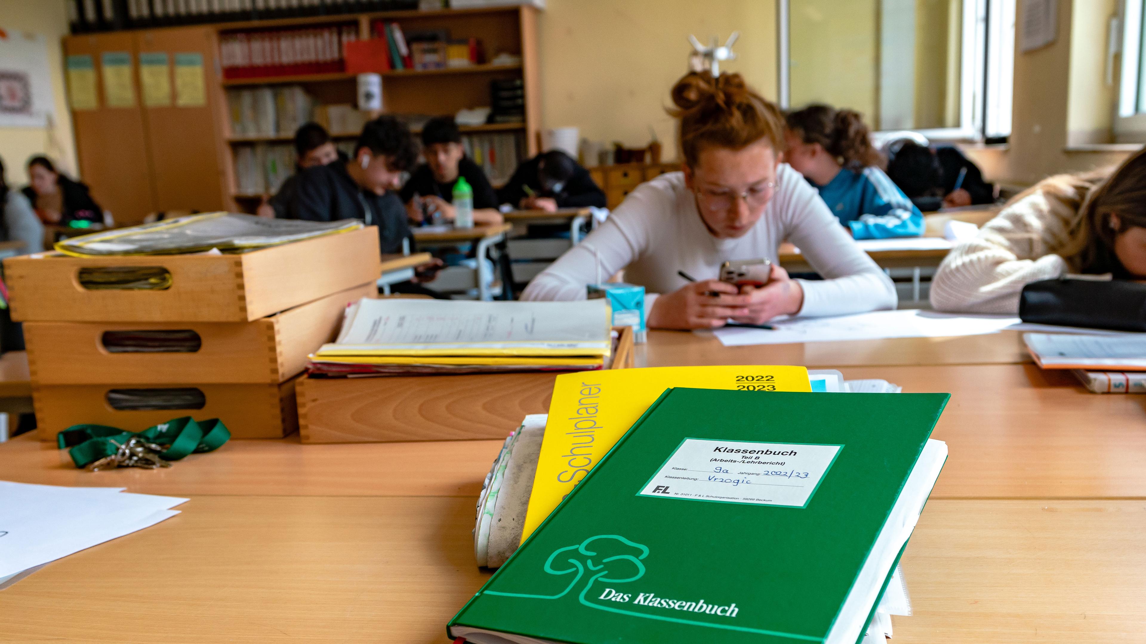Im Vordergrund liegt auf dem Lehrerpult das Klassenbuch der 9a. Im Hintergrund sitzen Schülerinnen und Schüler.