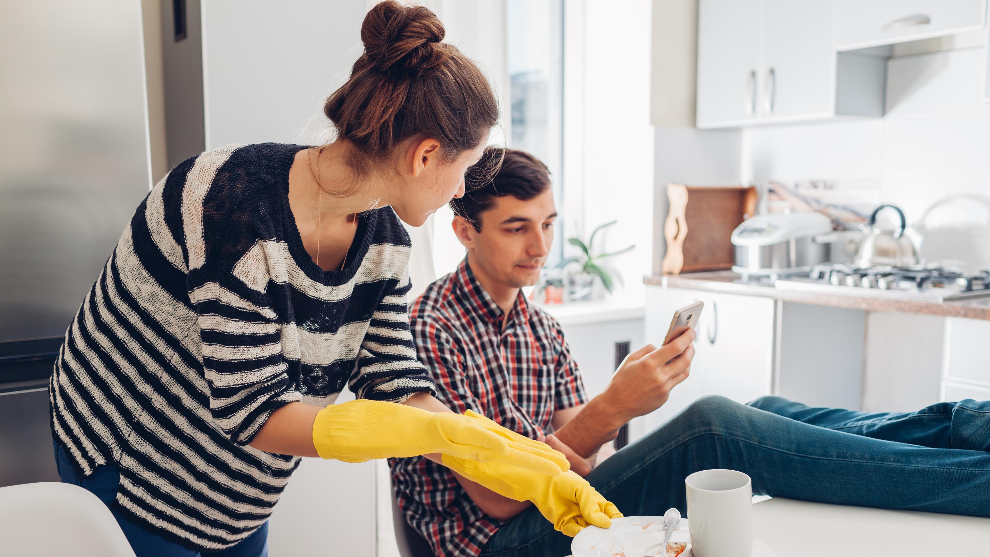 Eine Frau mit gelben Putzhandschuhen räumt dreckiges Geschirr ab, während ein Mann die Füße auf den Tisch gelegt hat und auf sein Handy schaut.